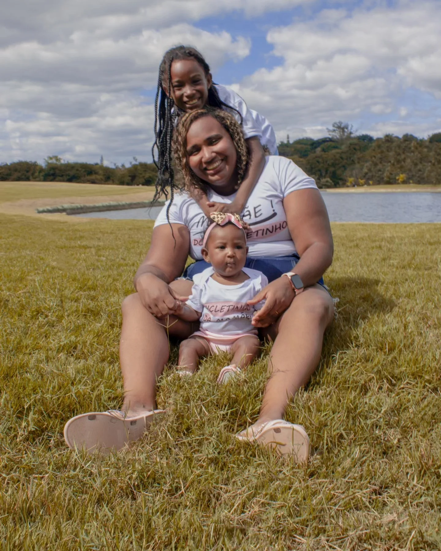 Ensaio em família ao ar livre à beira do lago com mãe sorrindo junto das suas duas filhas no gramado